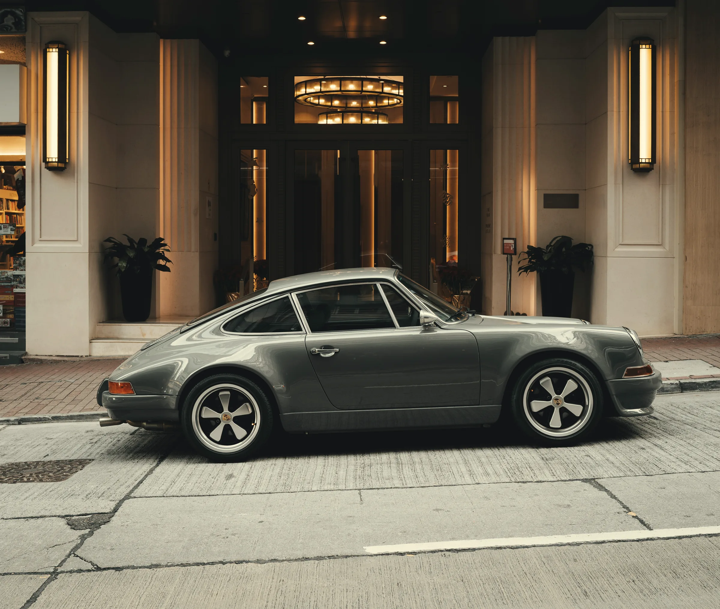 A grey classic rear-engined coupe parked side-on outside a lamplit hotel entrance